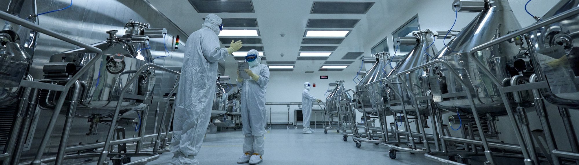 Cleanroom technicians in full protective gear working in a large pharmaceutical production room.