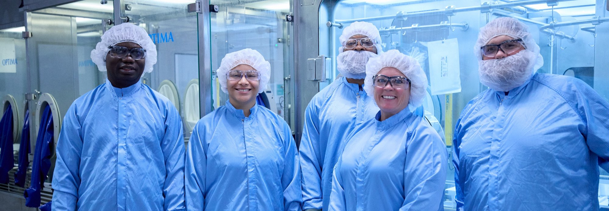 Diverse team of five laboratory technicians wearing blue cleanroom gear in a manufacturing facility.
